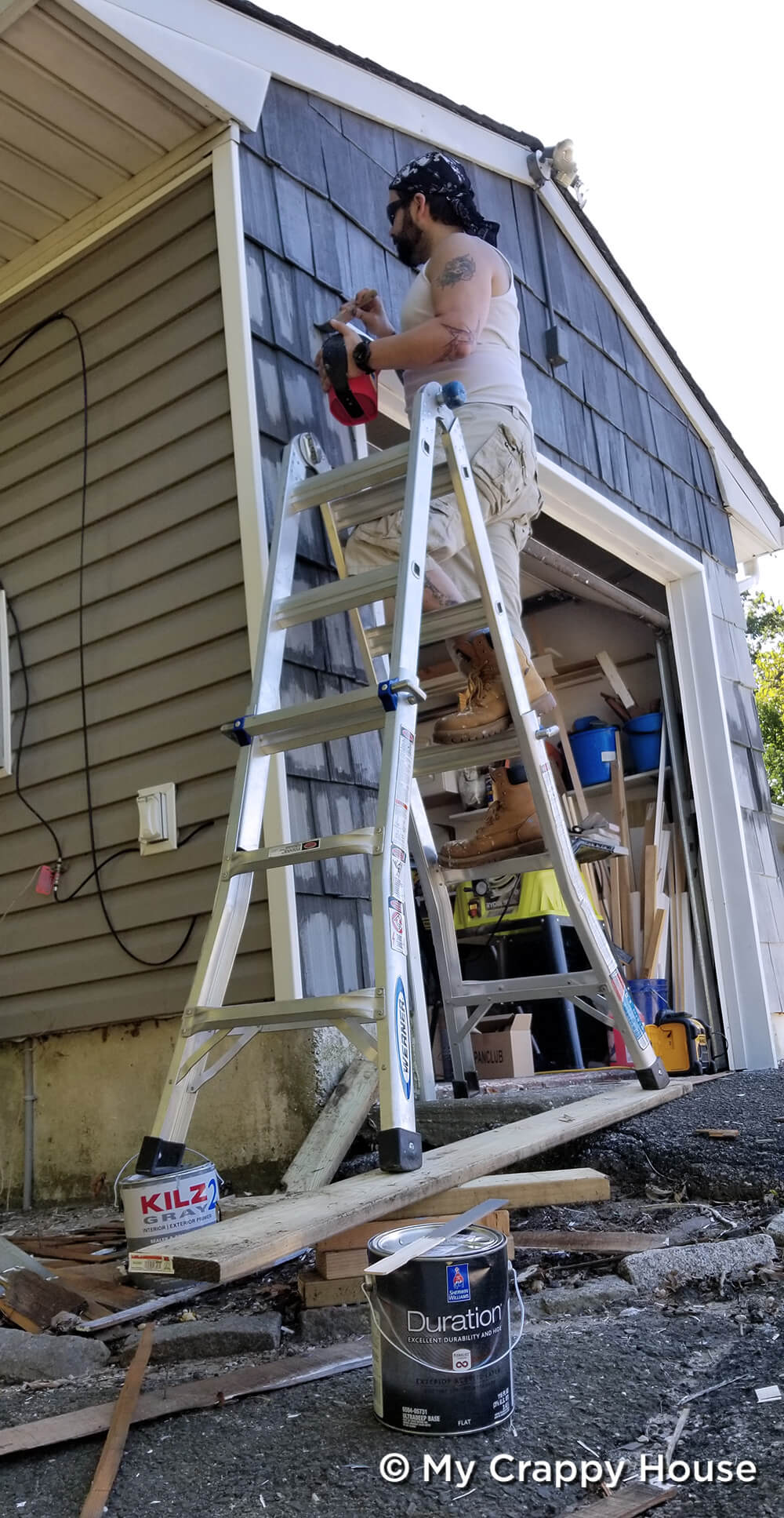 Man on precariously placed ladder painting cedar shingles gray on garage