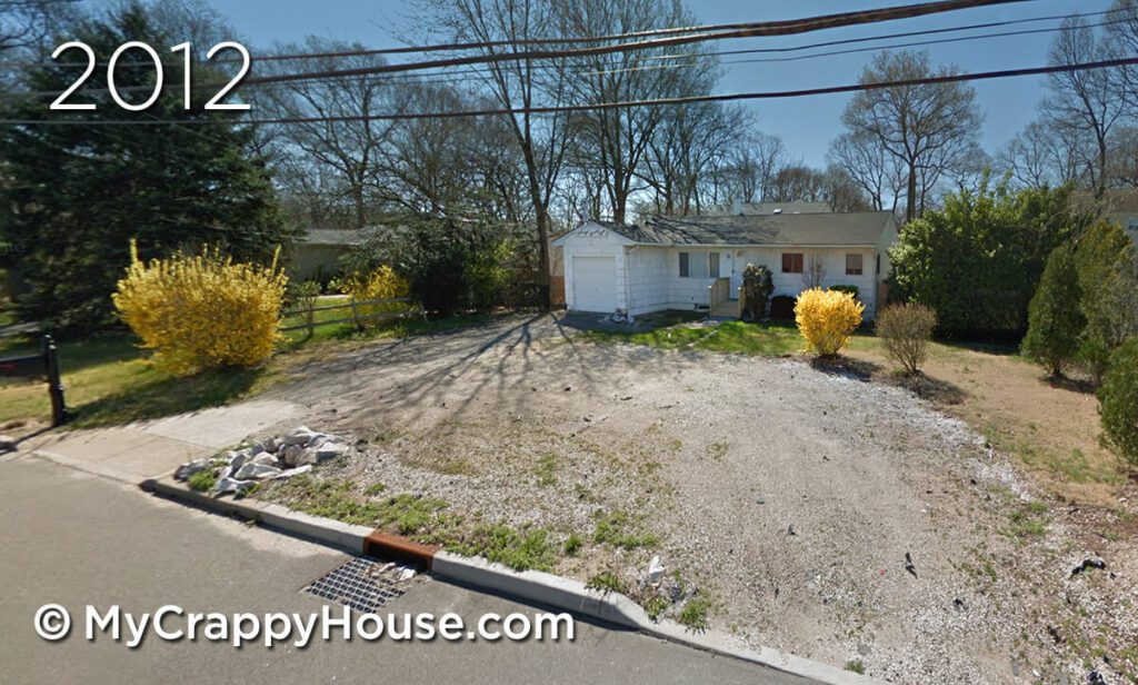 View from street of dingy white ranch house with a gravel and broken asphalt driveway