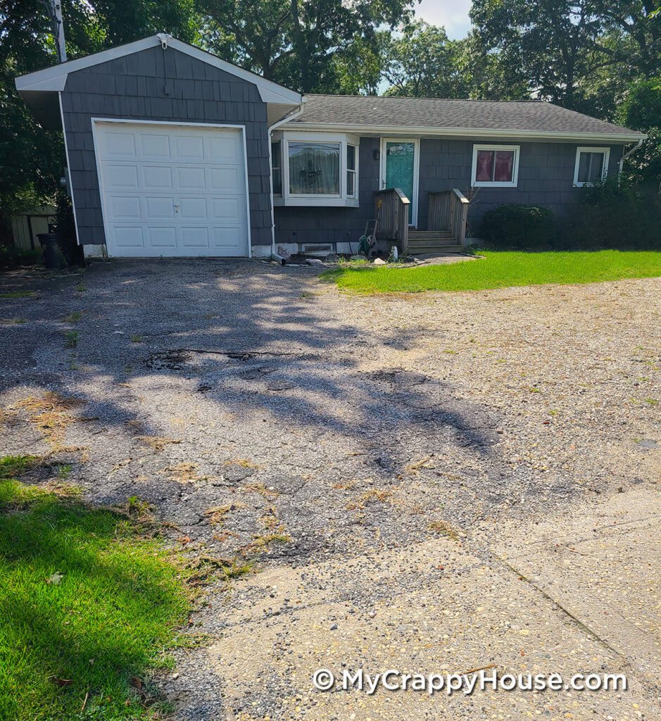 Gray house white trim with a very crappy driveway of cracked asphalt and rocks