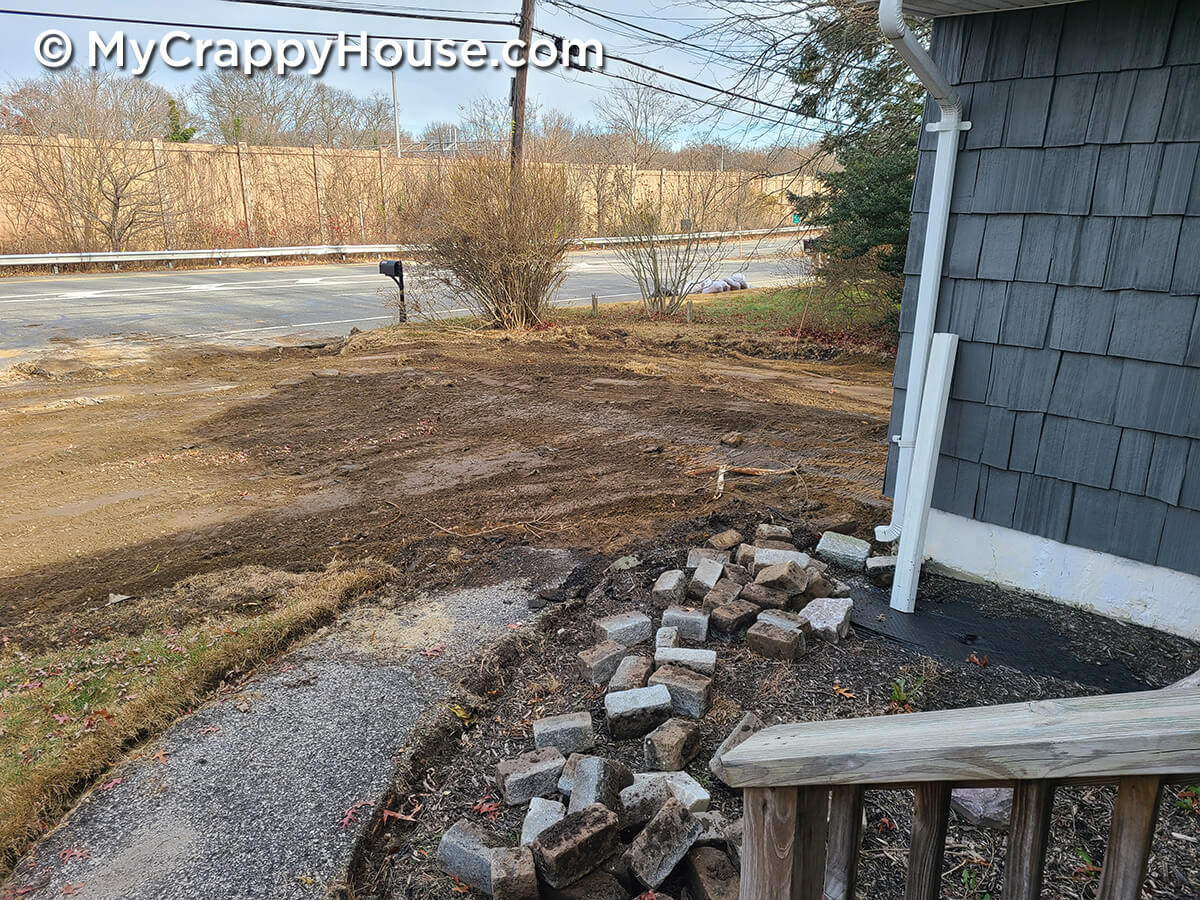 Dirt in front of gray house from removed driveway with pile of Belgian block next to old asphalt walkway