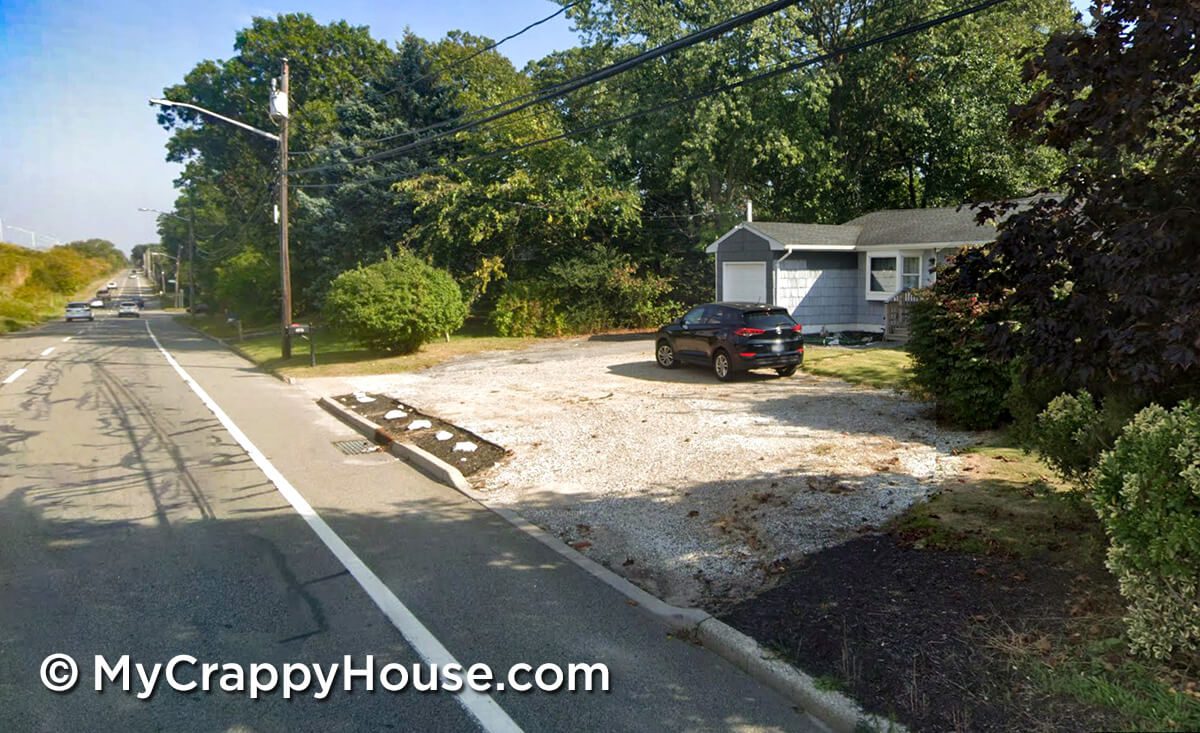 View from street of crappy gravel driveway with black car parked in front of half painted gray house