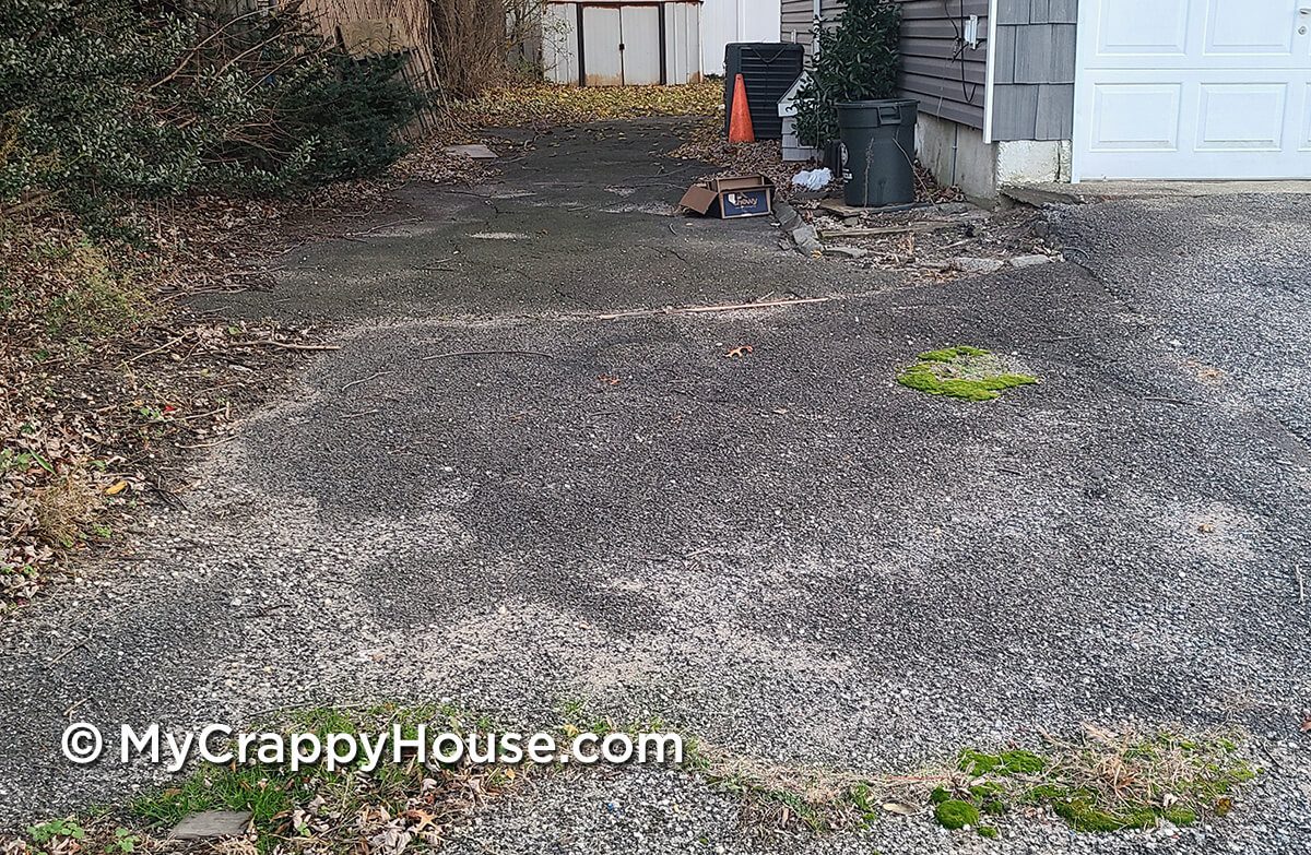 Broken asphalt driveway that goes along side a gray house