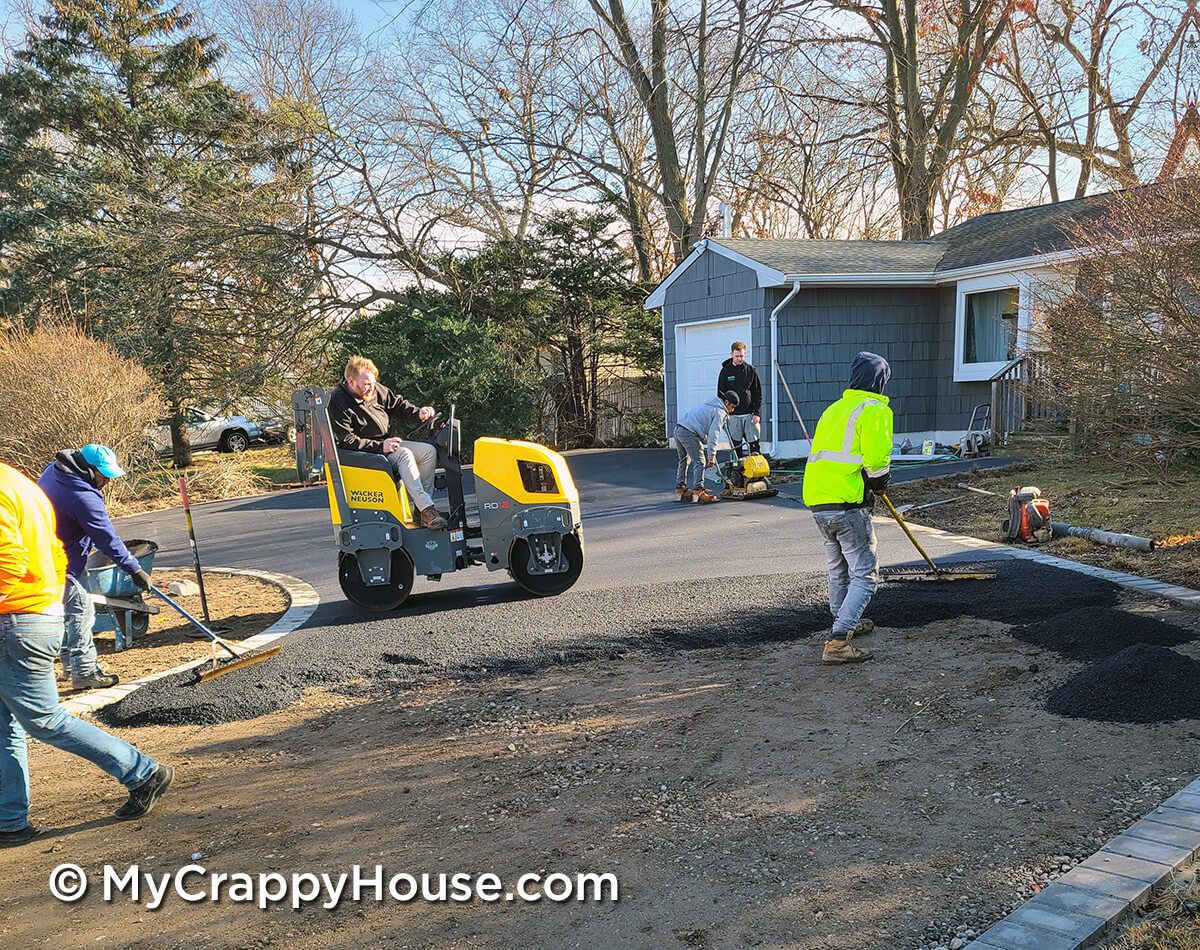 Man steamrolling asphalt on partially installed driveway. Several men raking asphalt.