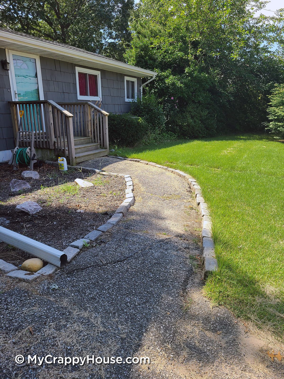 Gray ranch house with Tiffany blue door, wooden front steps, and uneven old asphalt walkway with Belgian block edges