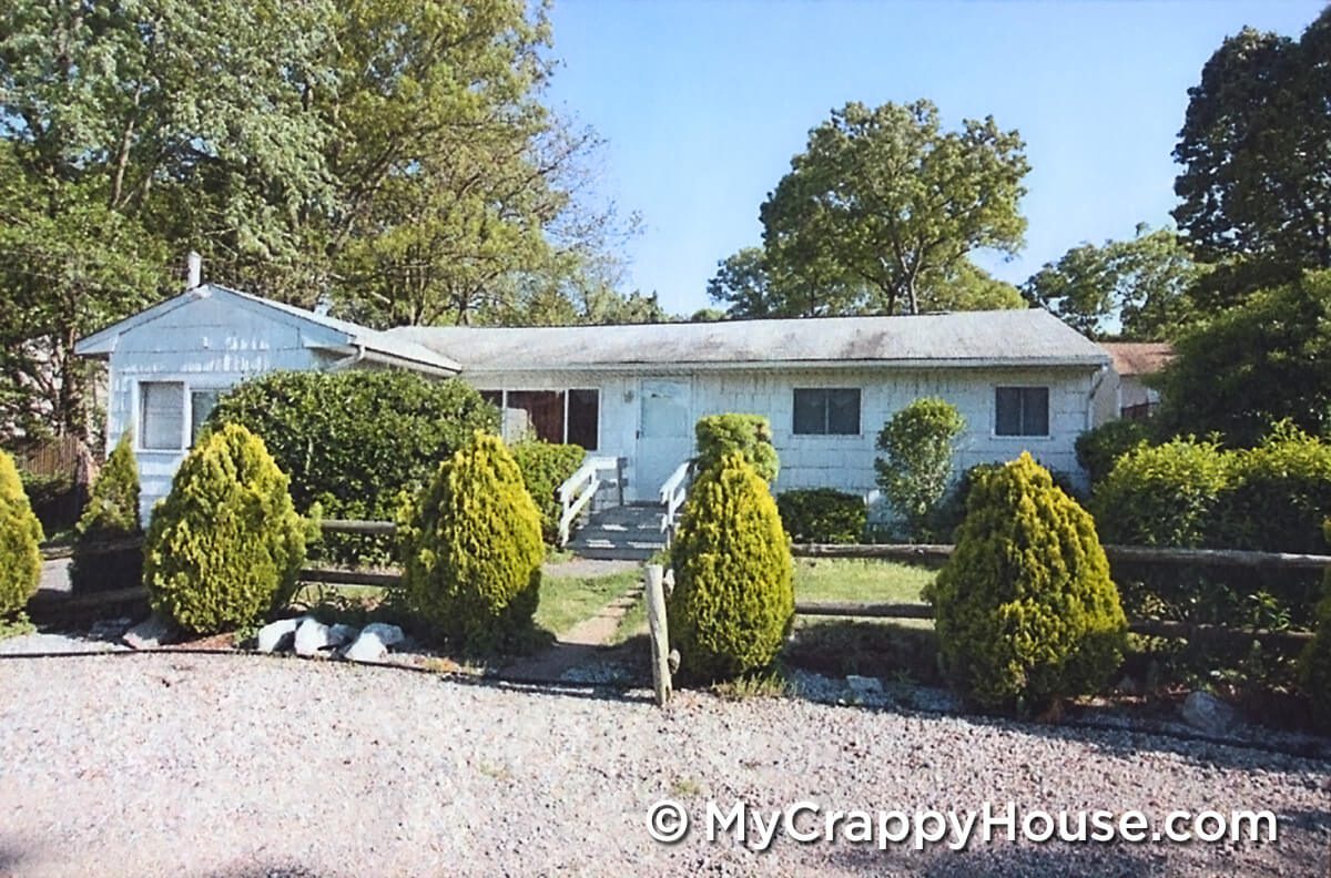 Run down white ranch house with arborvitae trees along a gravel driveway and a split rail fence