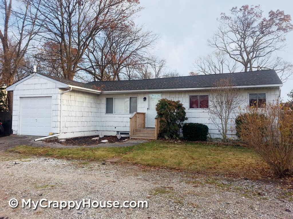 Unattractive white ranch house with poor landscaping and gravel driveway