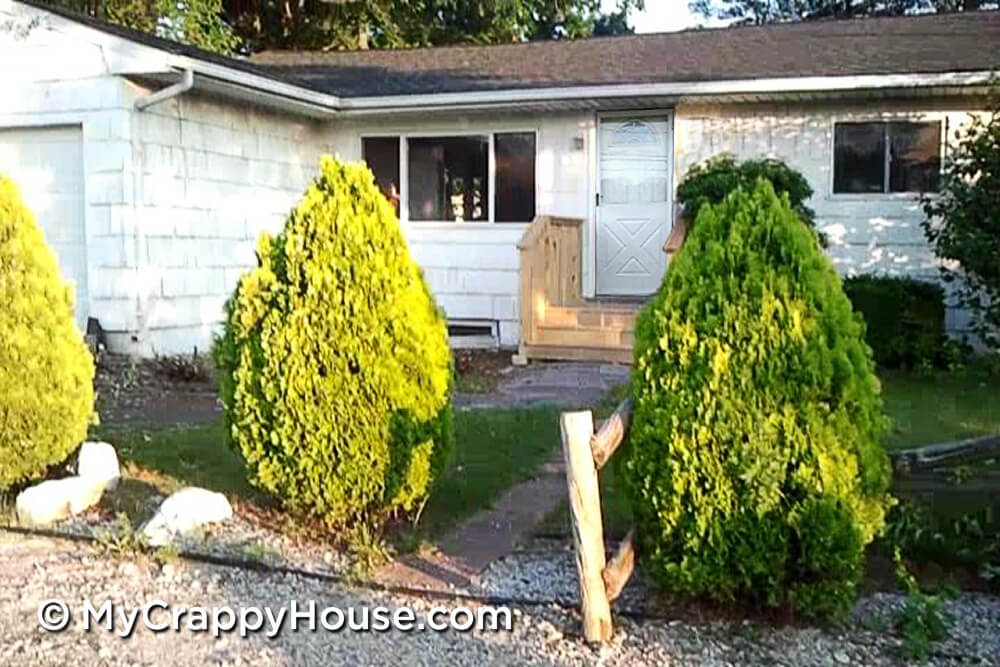 Run down white ranch house with arborvitae trees along a gravel driveway and a split rail fence