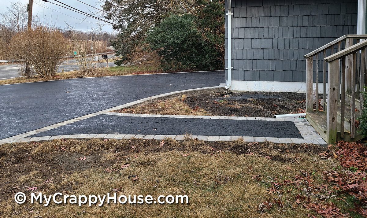 Straight asphalt walkway with flush recessed pavers leading to driveway in front of gray house