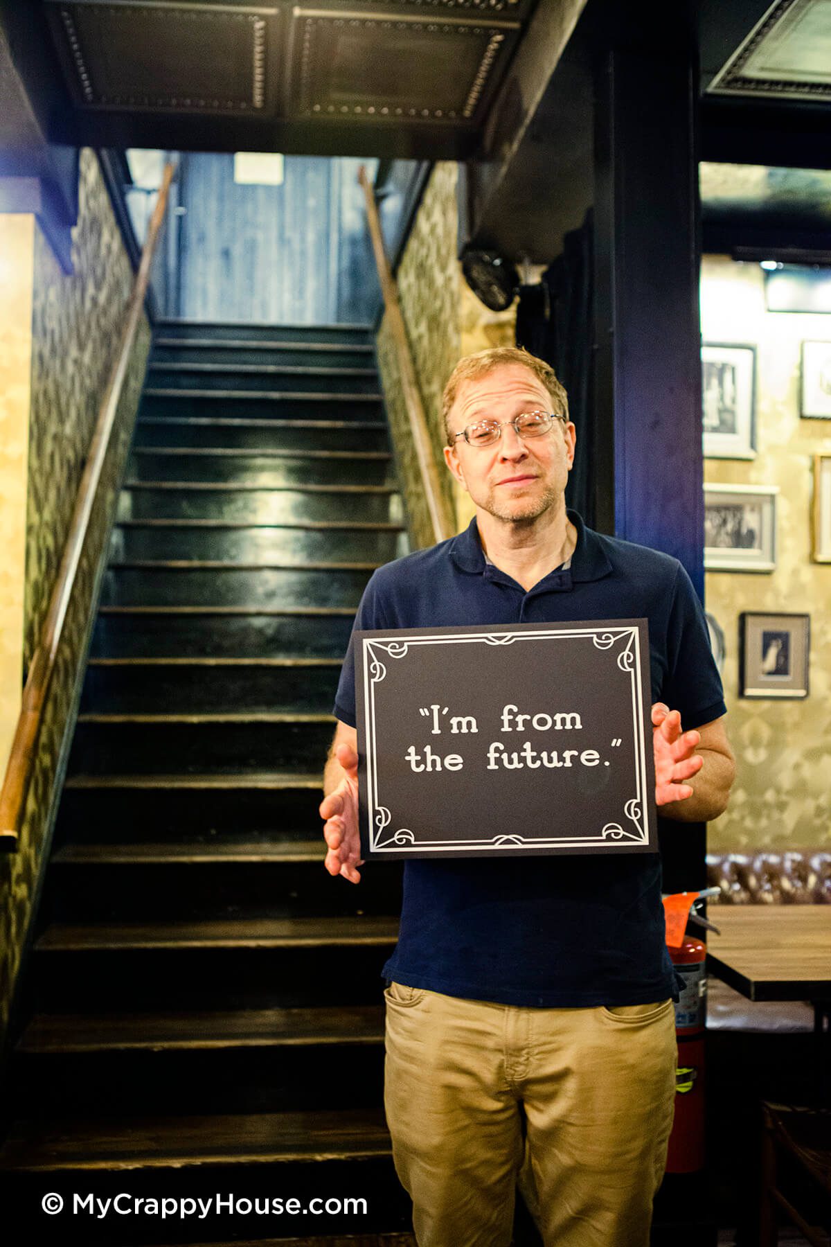 Man in modern clothes holding sign that says I'm from the future in front of staircase