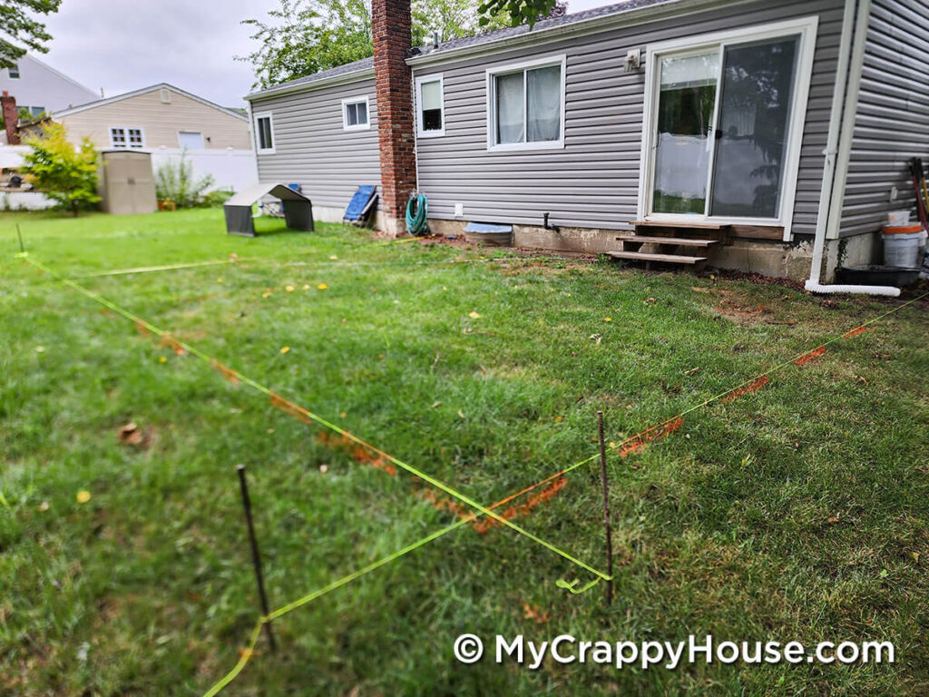 Backyard space marked with string layout lines showing the planned outline for a new deck behind a small gray house.