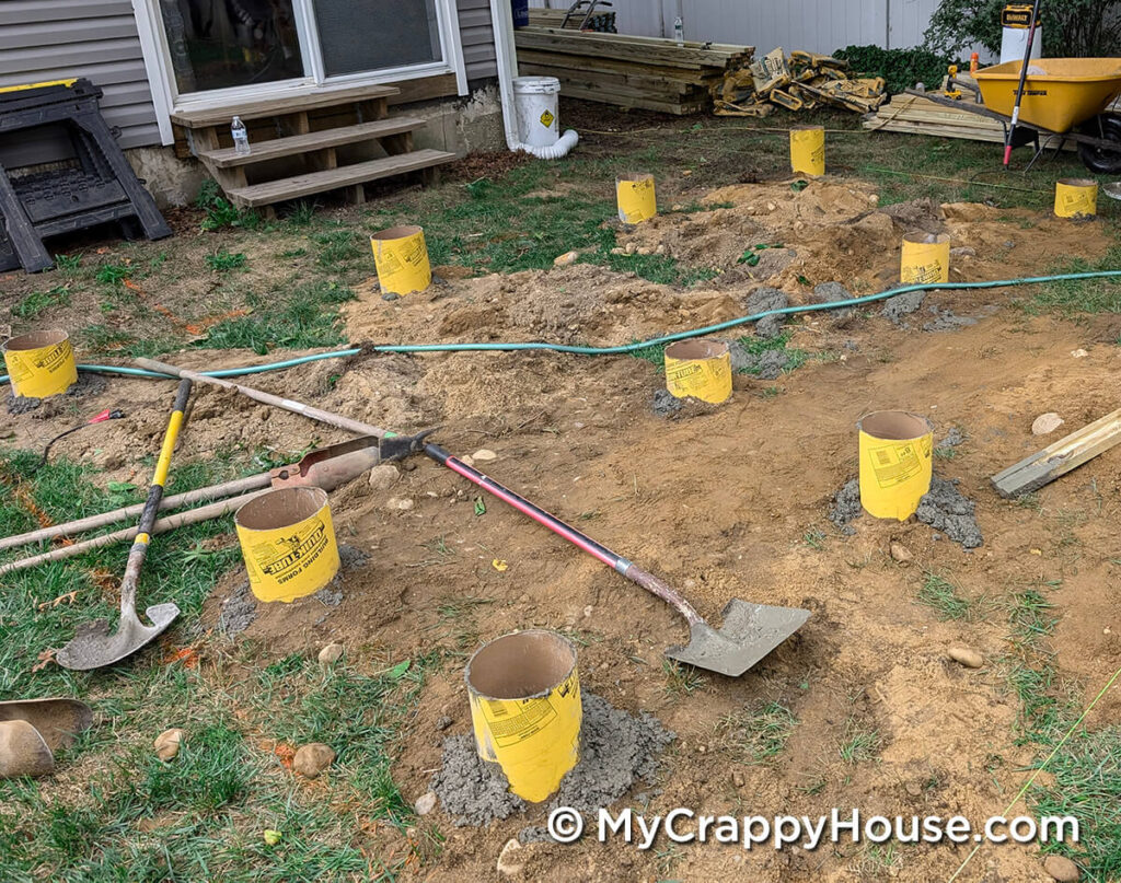 Freshly poured concrete deck footings in cardboard tubes across a dirt area with shovels and tools lying nearby.