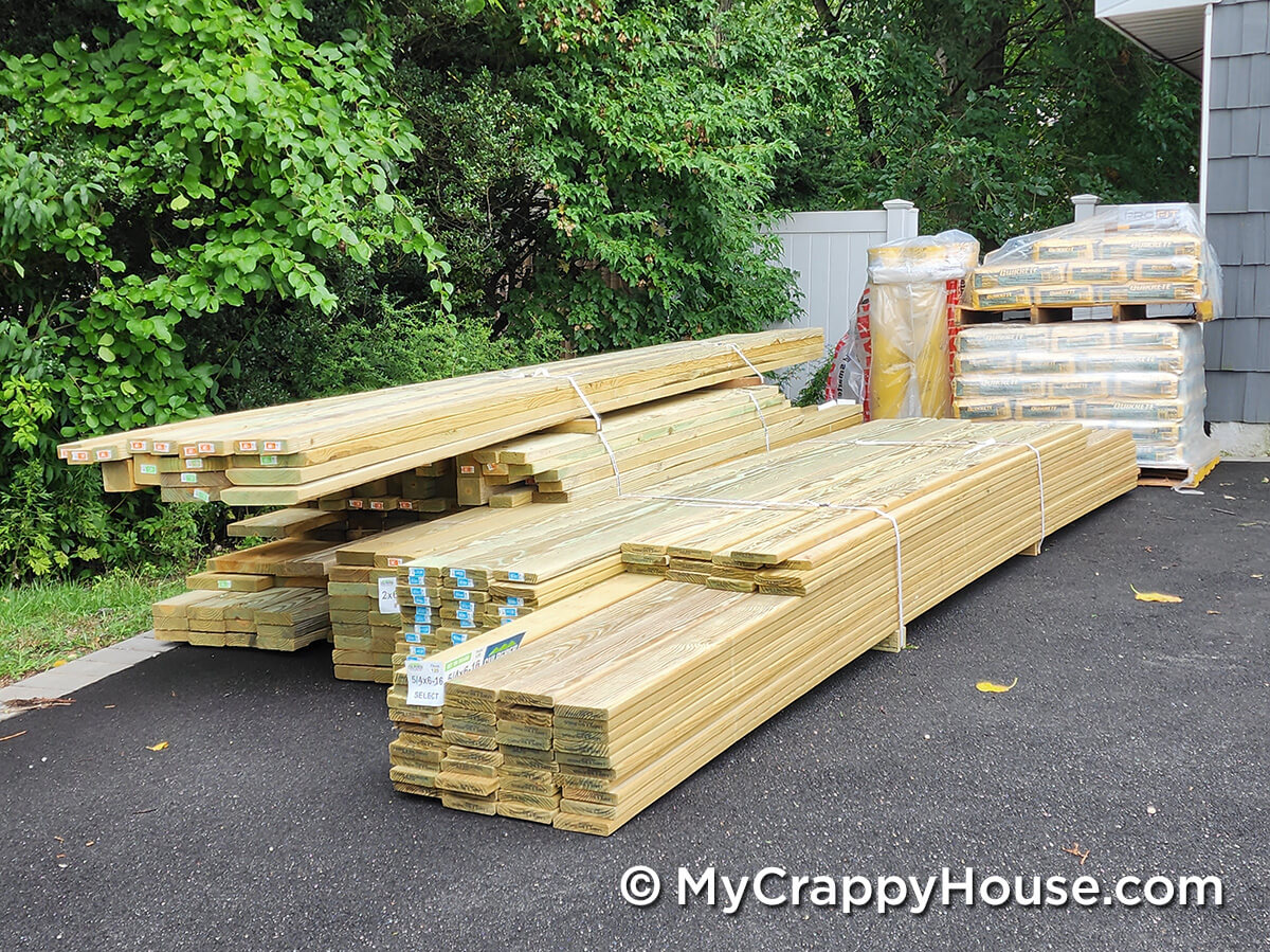 Large delivery of bundled pressure-treated lumber stacked on a driveway next to bags of concrete mix.