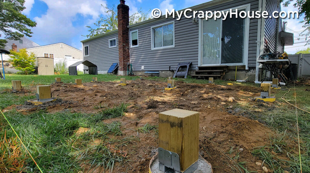Low-angle view of evenly spaced wooden deck posts anchored in concrete footings across a cleared dirt area behind a gray house.