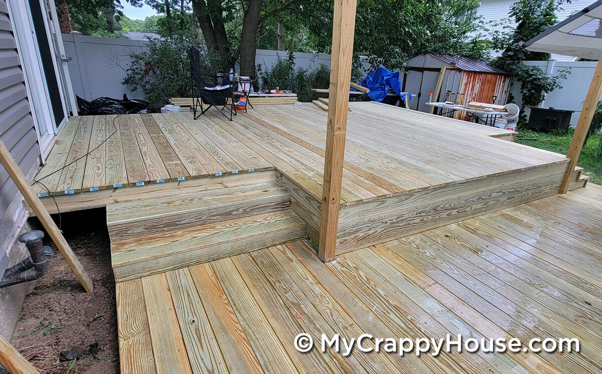 Back view of a newly built two-level wooden deck with fresh pressure-treated boards and temporary support posts, with tools and chairs scattered around the workspace.
