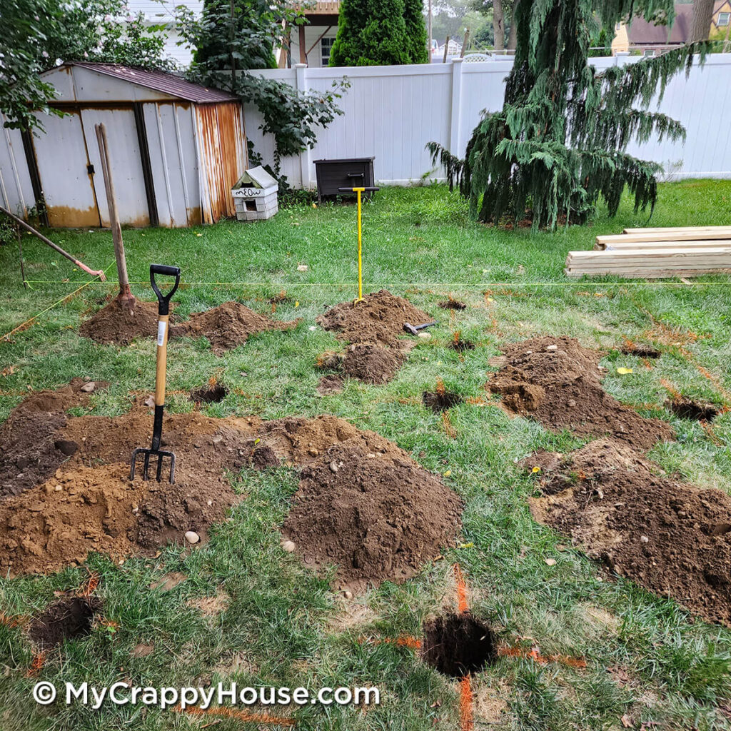 Rows of freshly dug post holes in a grassy backyard, with piles of soil, garden tools, and string layout lines marking the deck layout.
