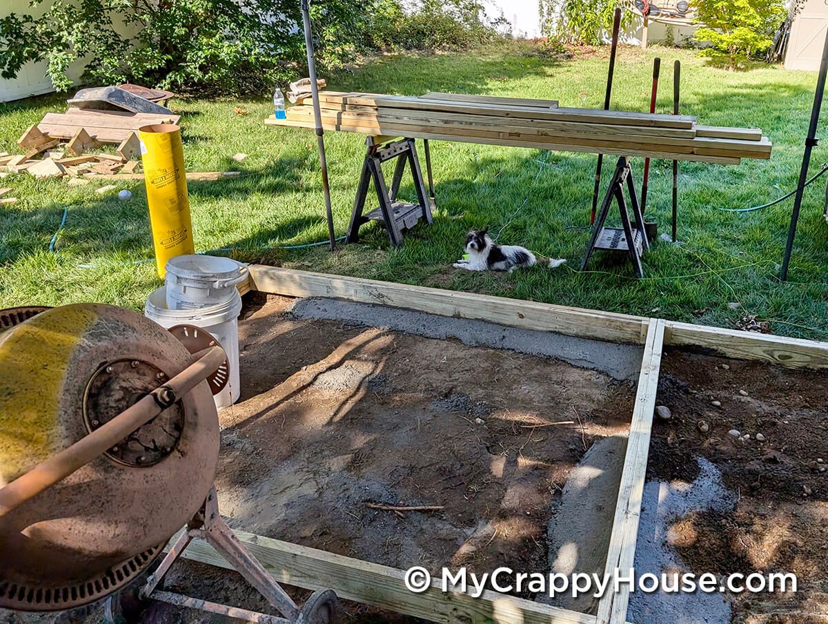Early deck construction area with a concrete mixer, buckets, lumber on sawhorses, and a small black-and-white dog resting in the grass nearby.