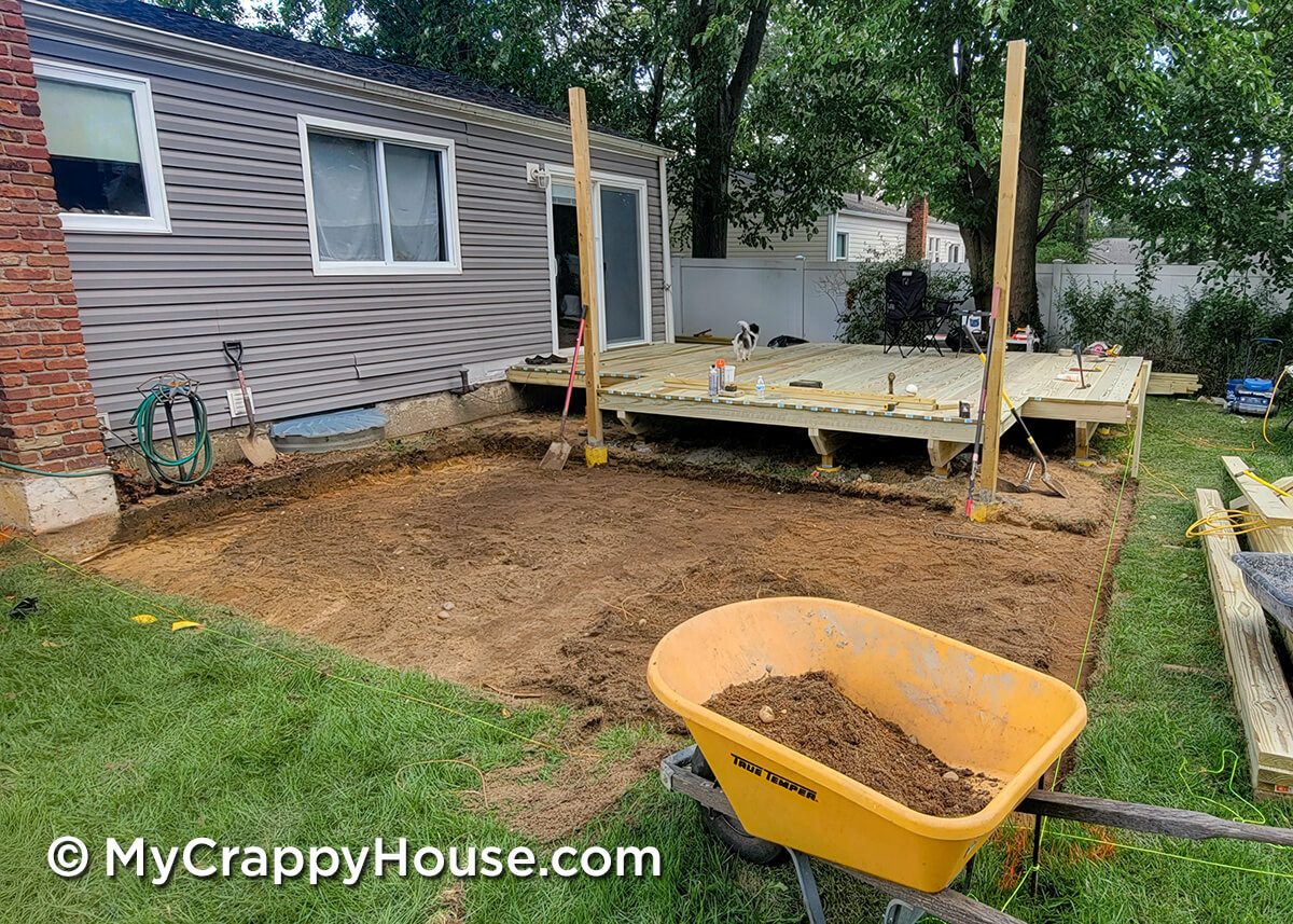 Cleared and leveled dirt area beside a house where a deck will be built, with a wheelbarrow, tools, and partially completed deck framing visible.