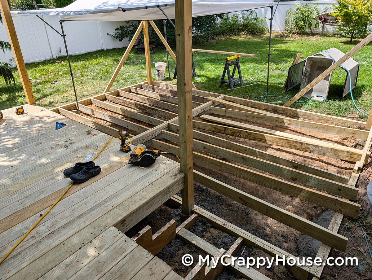 Partially built upper deck next to newly installed floor joists for a lower section, with tools and temporary shade canopy overhead.