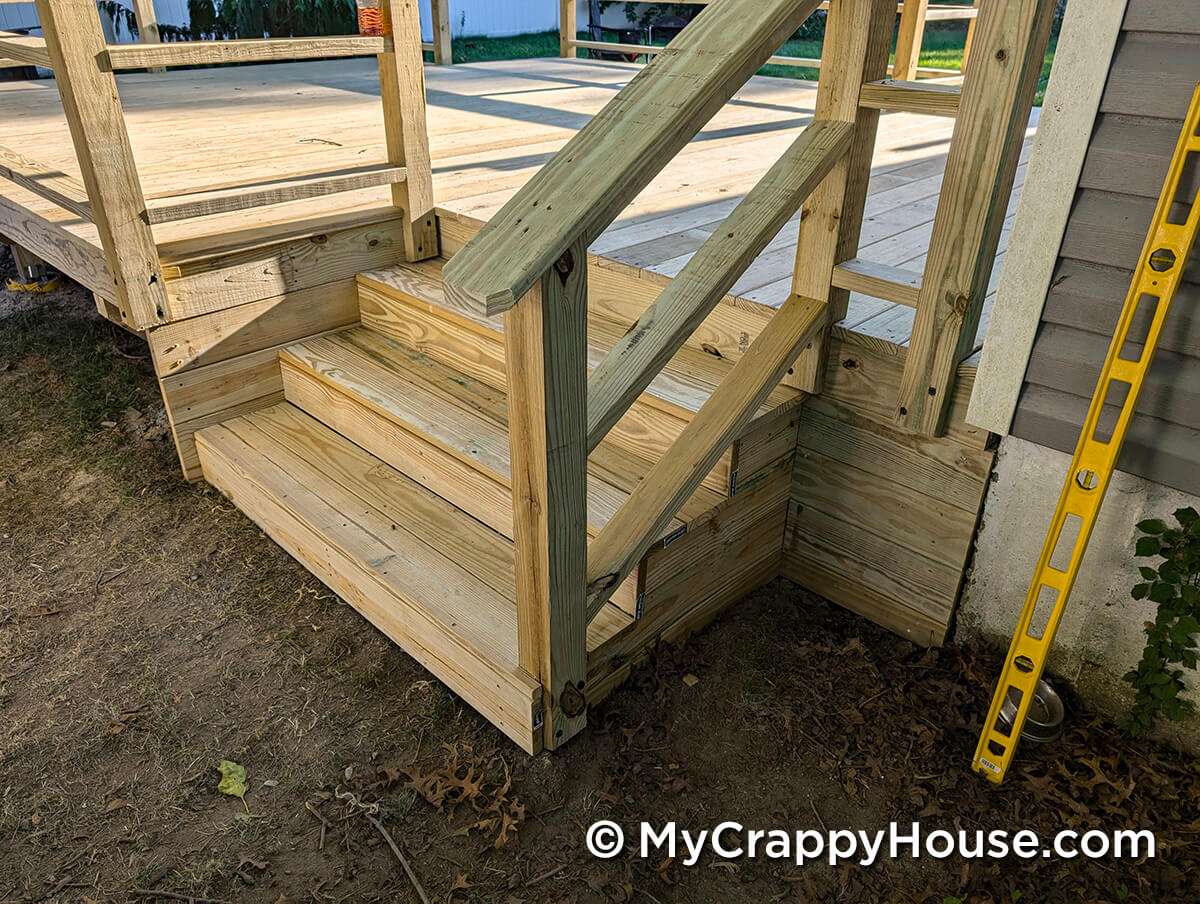 Newly constructed wooden deck stairs with partially built railing, set next to the house with a level tool leaning against the wall.