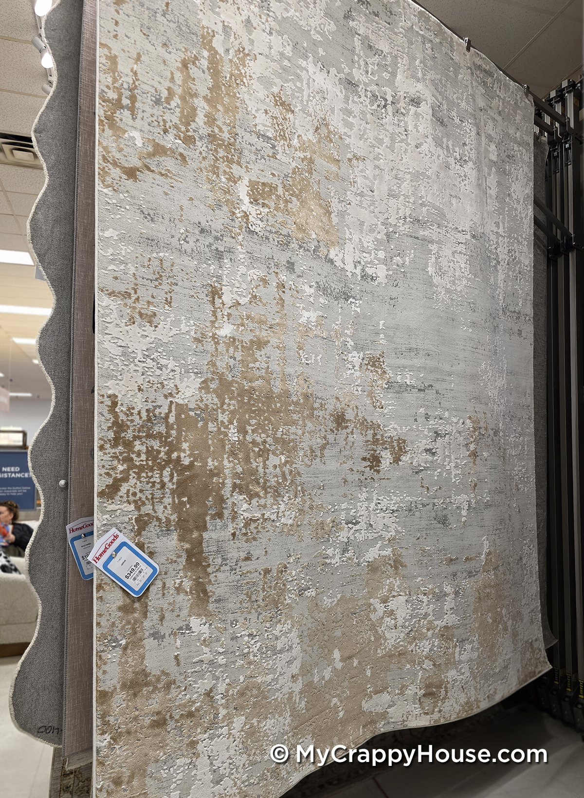 Large area rug on display in a store, featuring an abstract textured pattern in beige, gray, gold, and cream tones, shown hanging from a rug rack with price tags attached