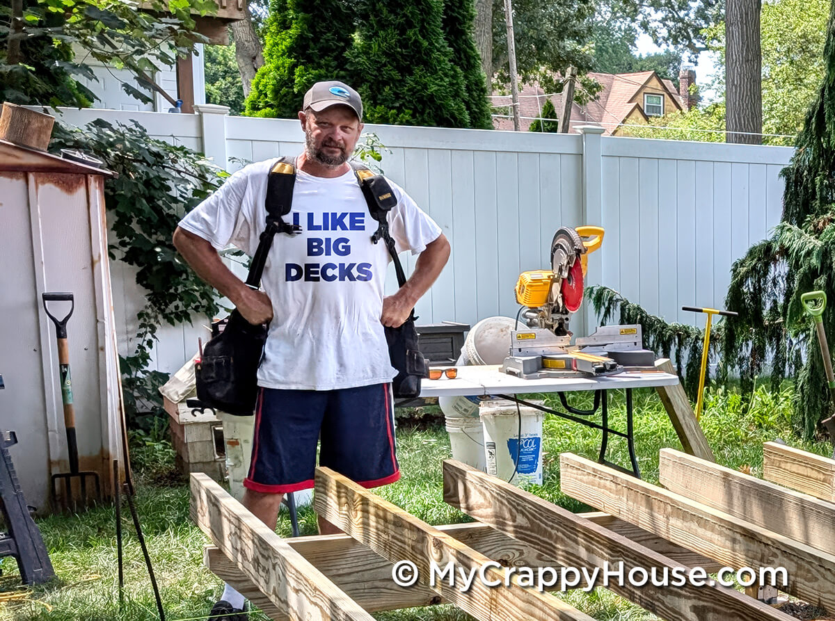 Man standing at a backyard construction site wearing a tool belt and a T-shirt that says I LIKE BIG DECKS, with lumber, tools, and a miter saw visible around him.