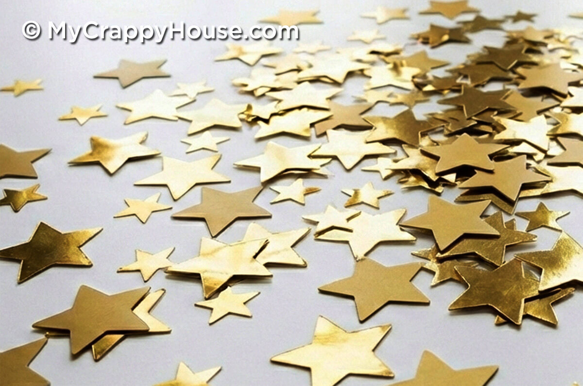 Close-up photograph of scattered gold star-shaped confetti on a white surface, with stars in varying sizes overlapping and reflecting light.
