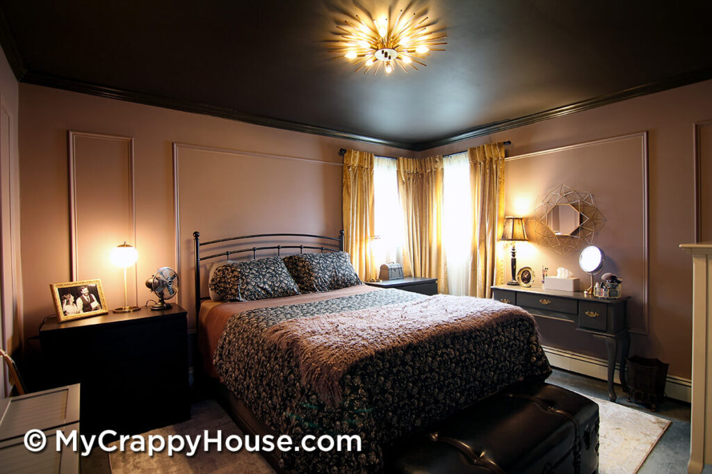 Finished bedroom renovation with dark painted ceiling, mauve walls with panel molding, black metal bed, layered bedding, and gold starburst ceiling light, warmly lit with bedside lamps and curtains framing corner windows.