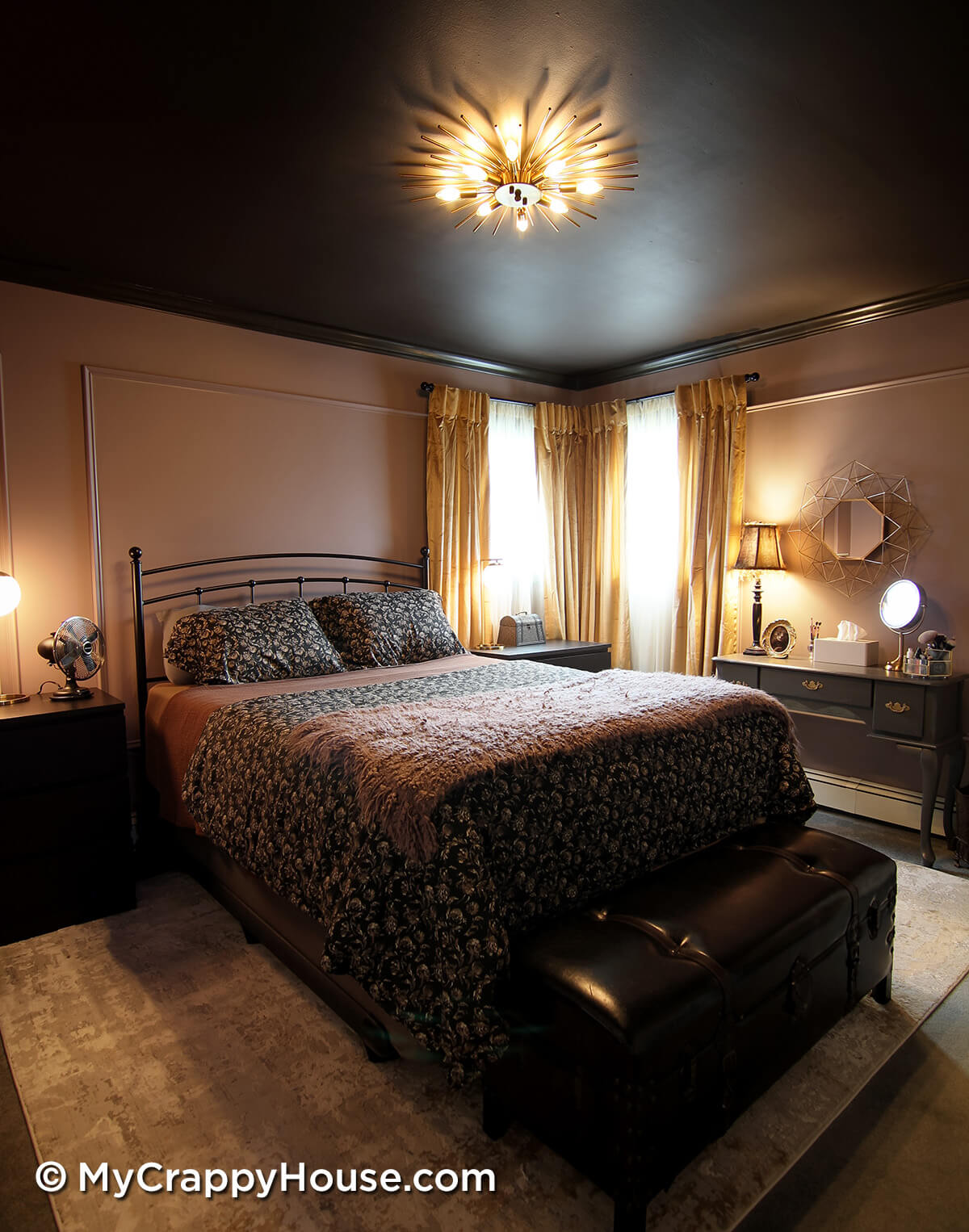 Finished bedroom renovation with dark painted ceiling, mauve walls with panel molding, black metal bed, layered bedding, and gold starburst ceiling light, warmly lit with bedside lamps and curtains framing corner windows.