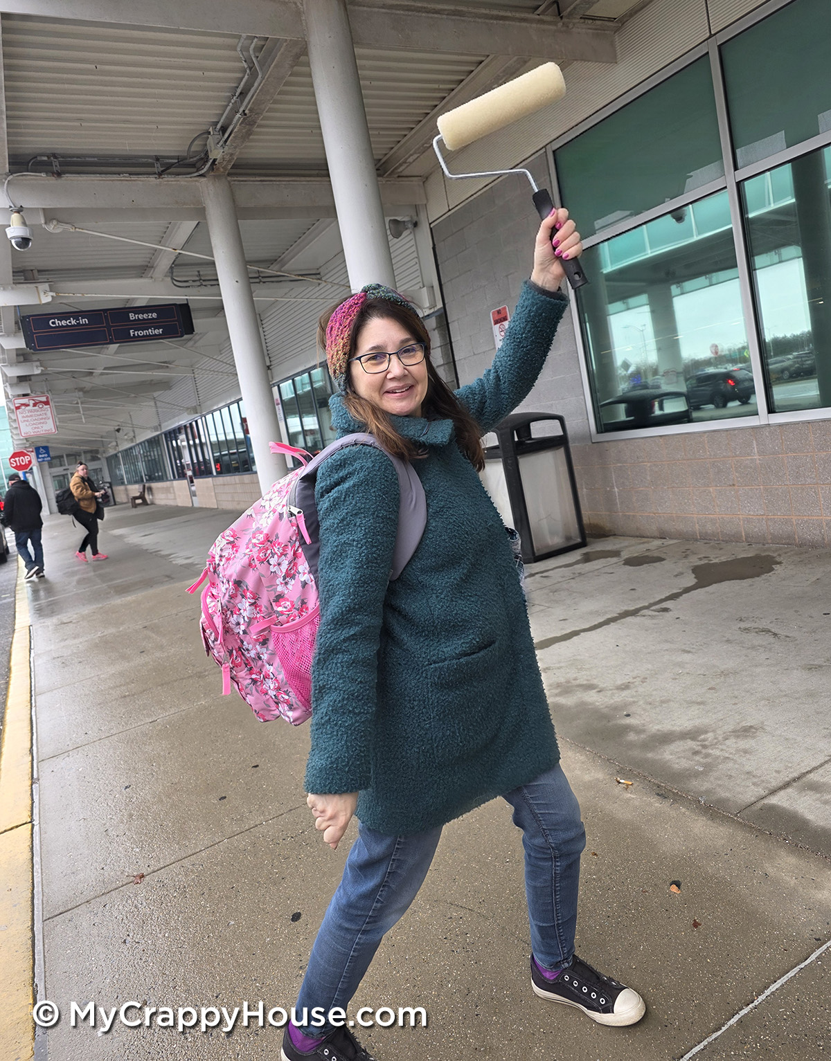 Woman outside an airport terminal holding a paint roller and wearing a backpack, smiling before leaving for a DIY bedroom renovation project.