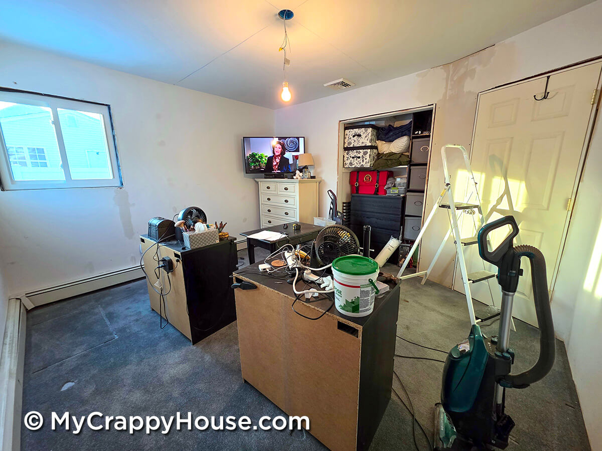 Cluttered unfinished bedroom with exposed light bulb, scattered tools and cords, a TV on a dresser, and a partially filled open closet during renovation.