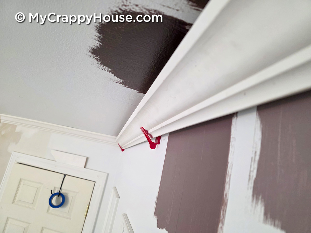 Close-up of crown molding near the ceiling ready to be installed with dark paint color swatches being tested on the wall during a bedroom makeover.