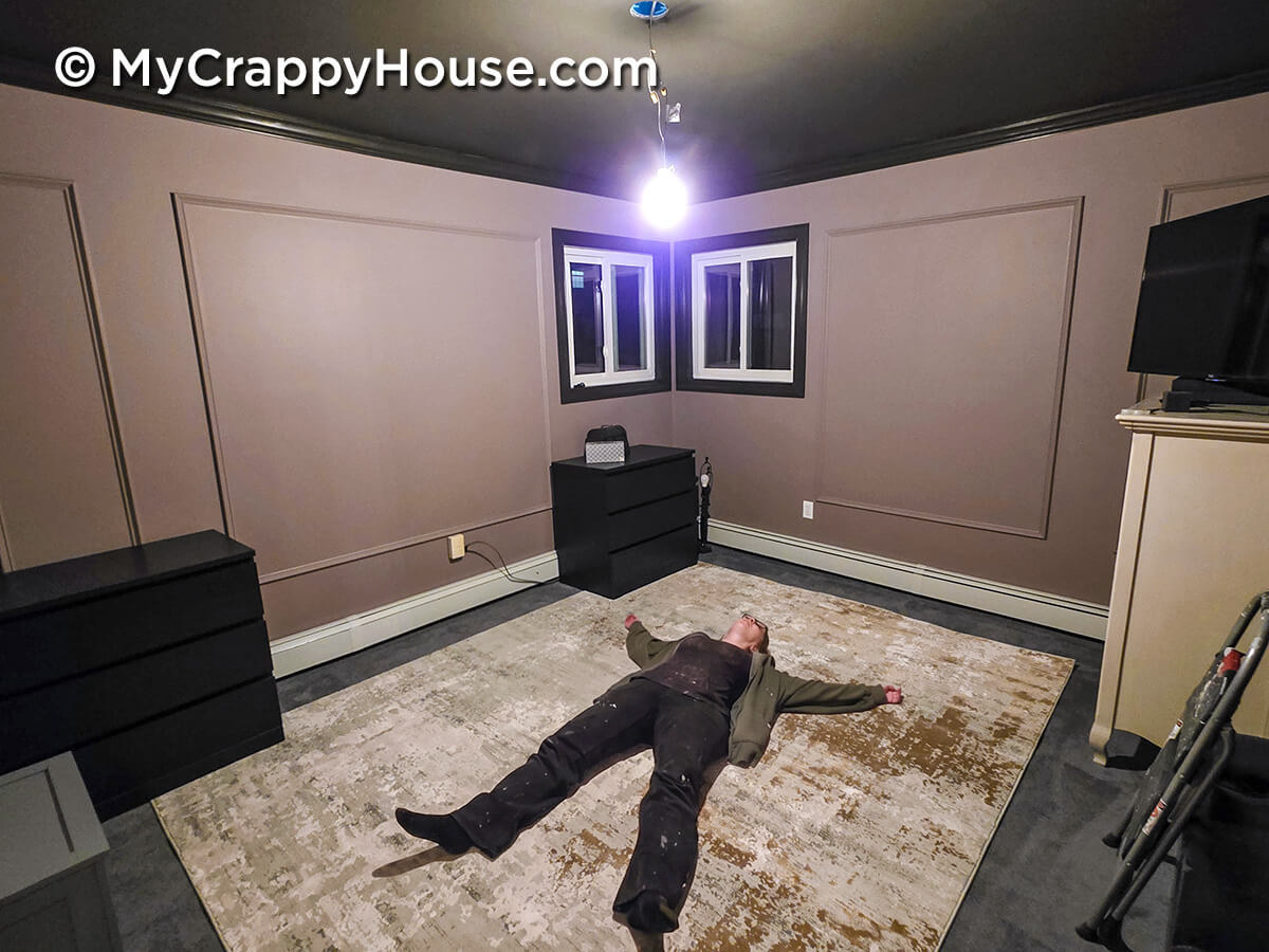 Completed bedroom renovation with dark ceiling, mauve walls, and decorative wall molding, showing person lying on floor exhausted after DIY project