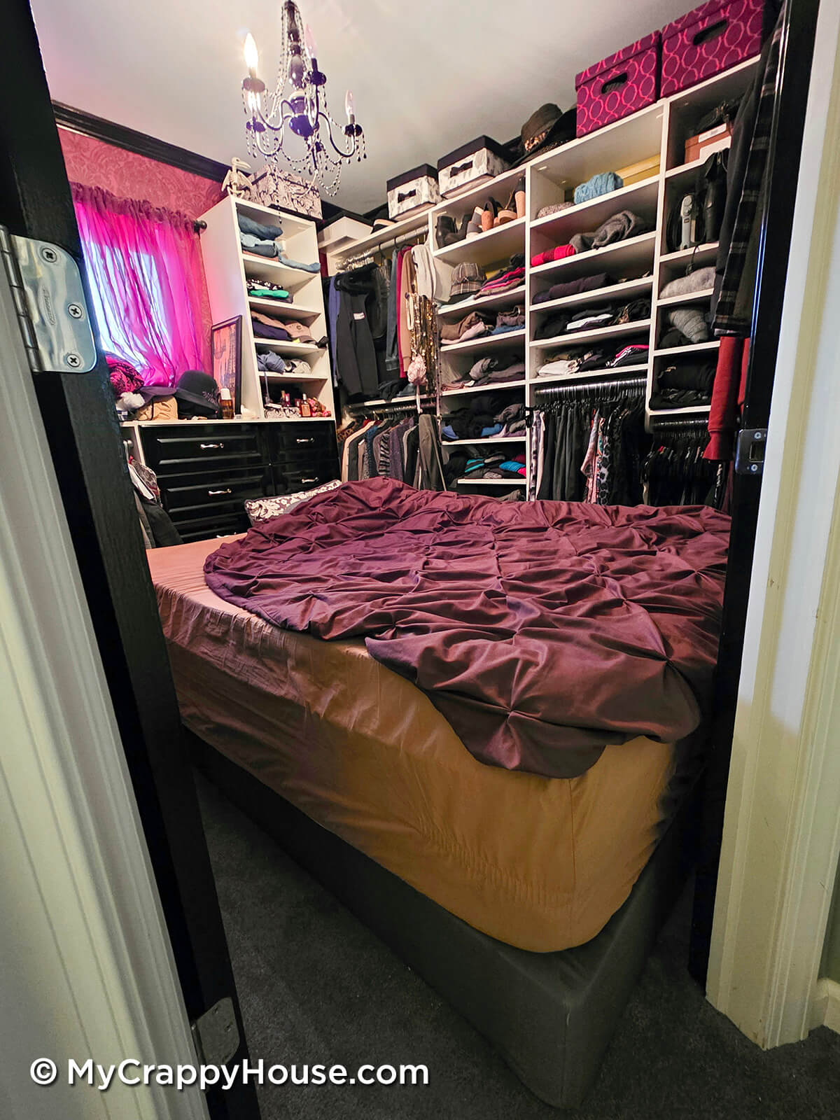 Small bedroom converted into a walk-in closet with built-in shelving full of clothes, a bed squeezed into the center, and a chandelier overhead.