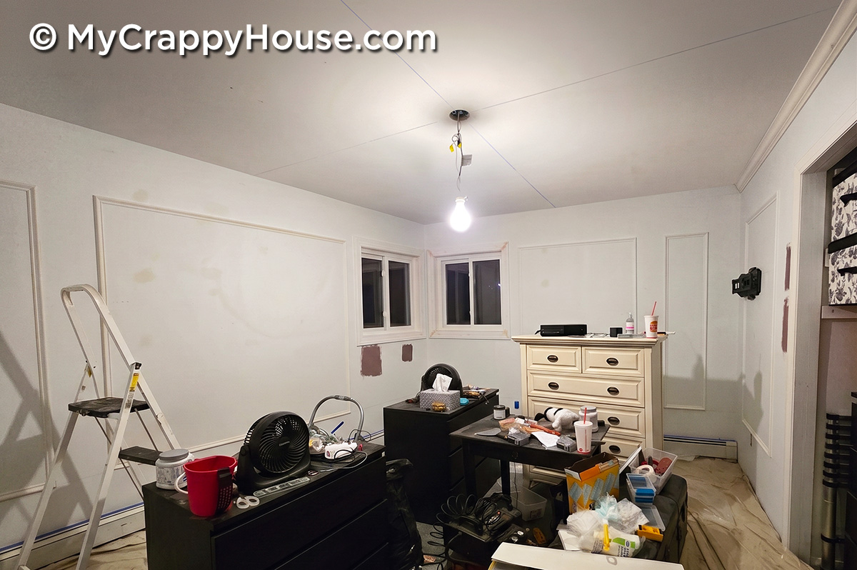 Partially finished bedroom renovation with wall panel molding installed, tools and supplies scattered around, and an exposed light bulb hanging from the ceiling.