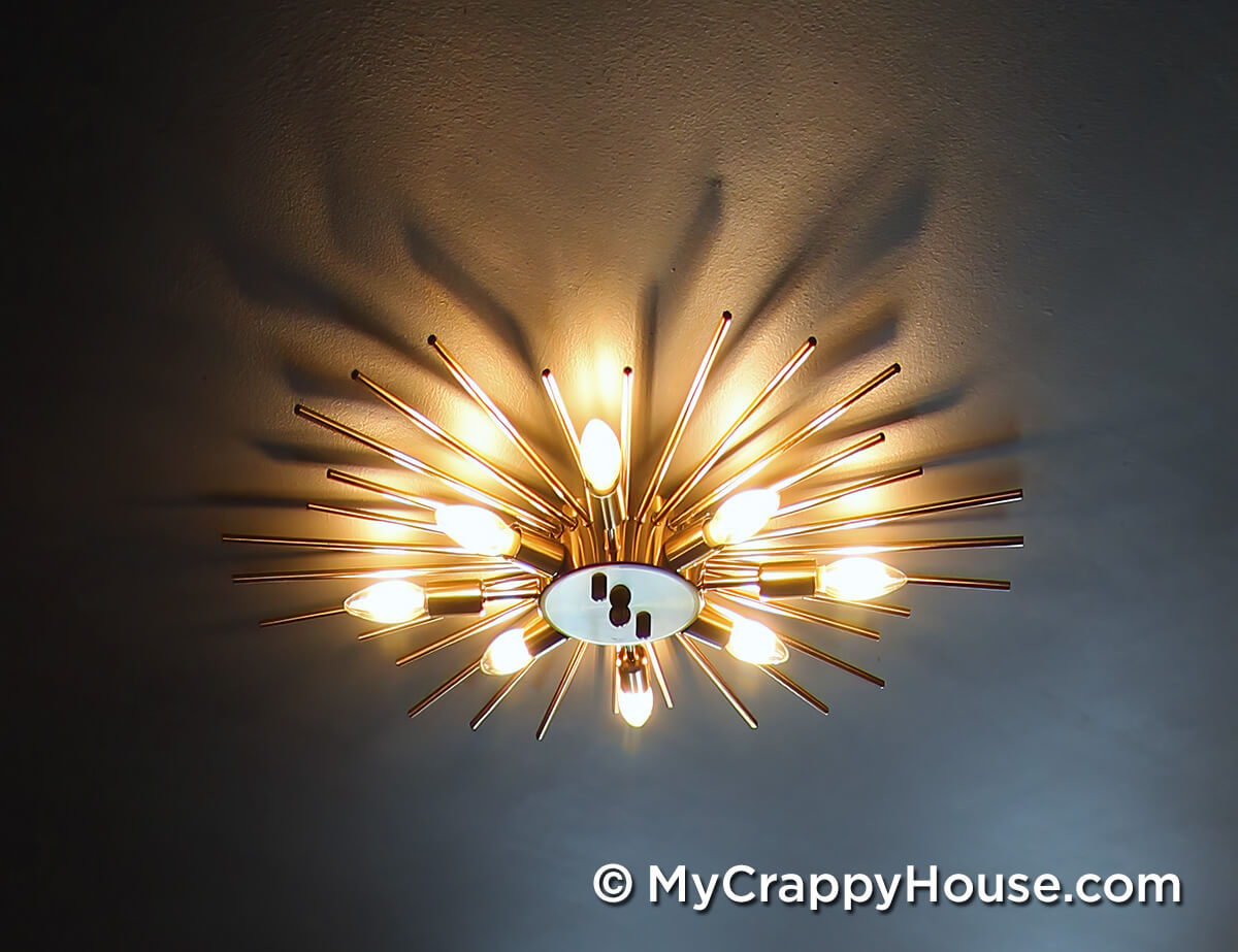 Gold starburst ceiling light fixture with exposed bulbs casting warm light and shadows across a dark painted ceiling in finished bedroom.