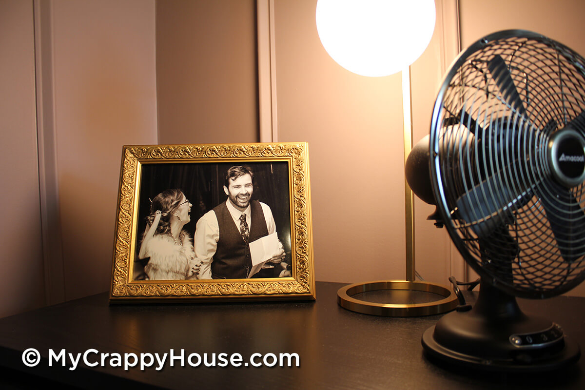 Nightstand styling with framed photo, round globe lamp, and small black fan on a dark dresser against mauve paneled wall in finished bedroom.