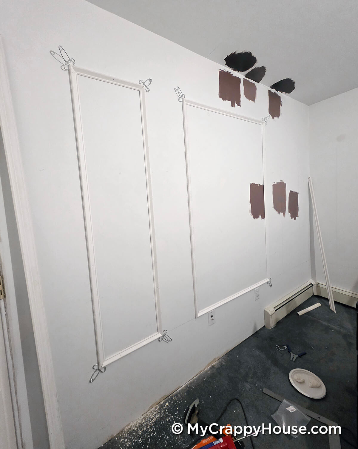 Wall with newly installed panel molding frames held in place by spring clamps, with paint color swatches on the wall and ceiling and tools scattered on the floor during bedroom renovation.
