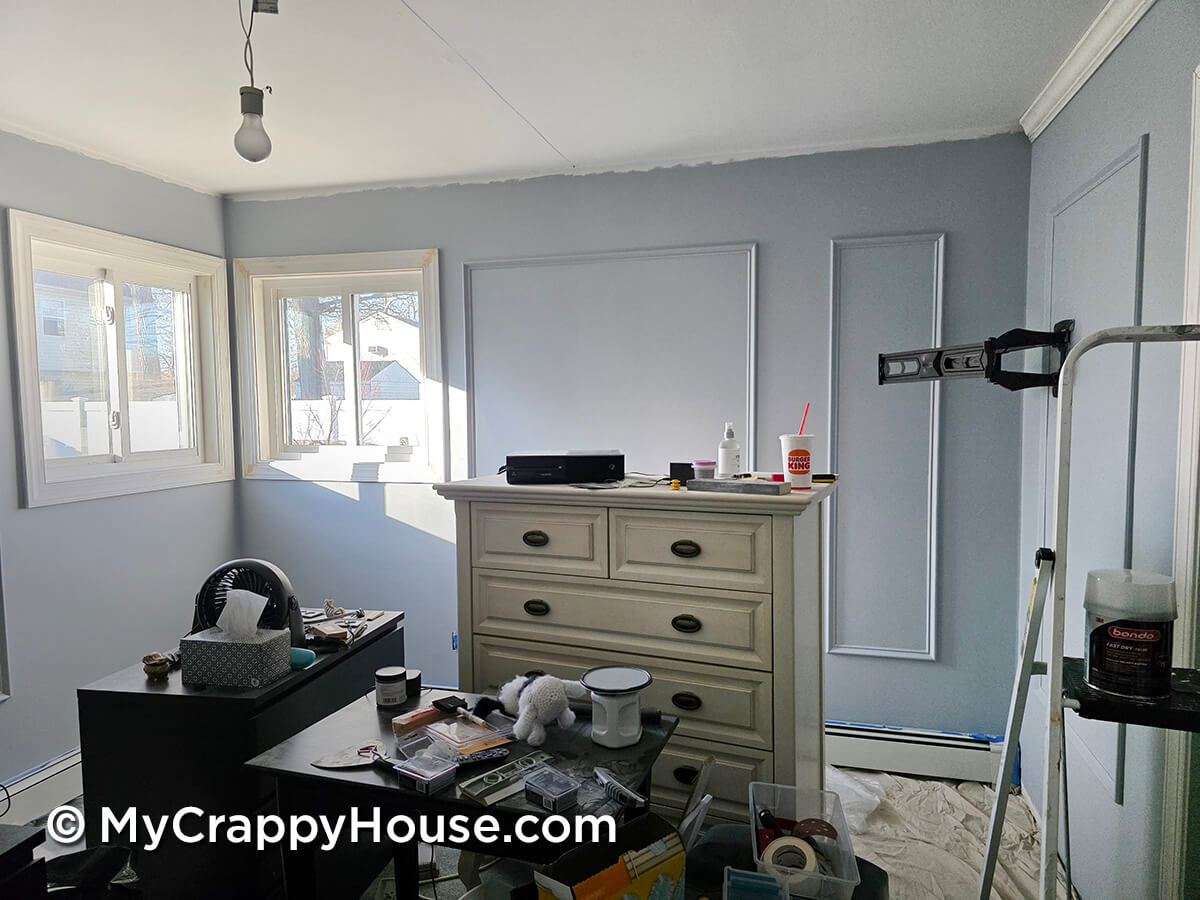 DIY bedroom renovation with fresh primer on the walls, wall panel molding installed, and room cluttered with tools and supplies mid-makeover