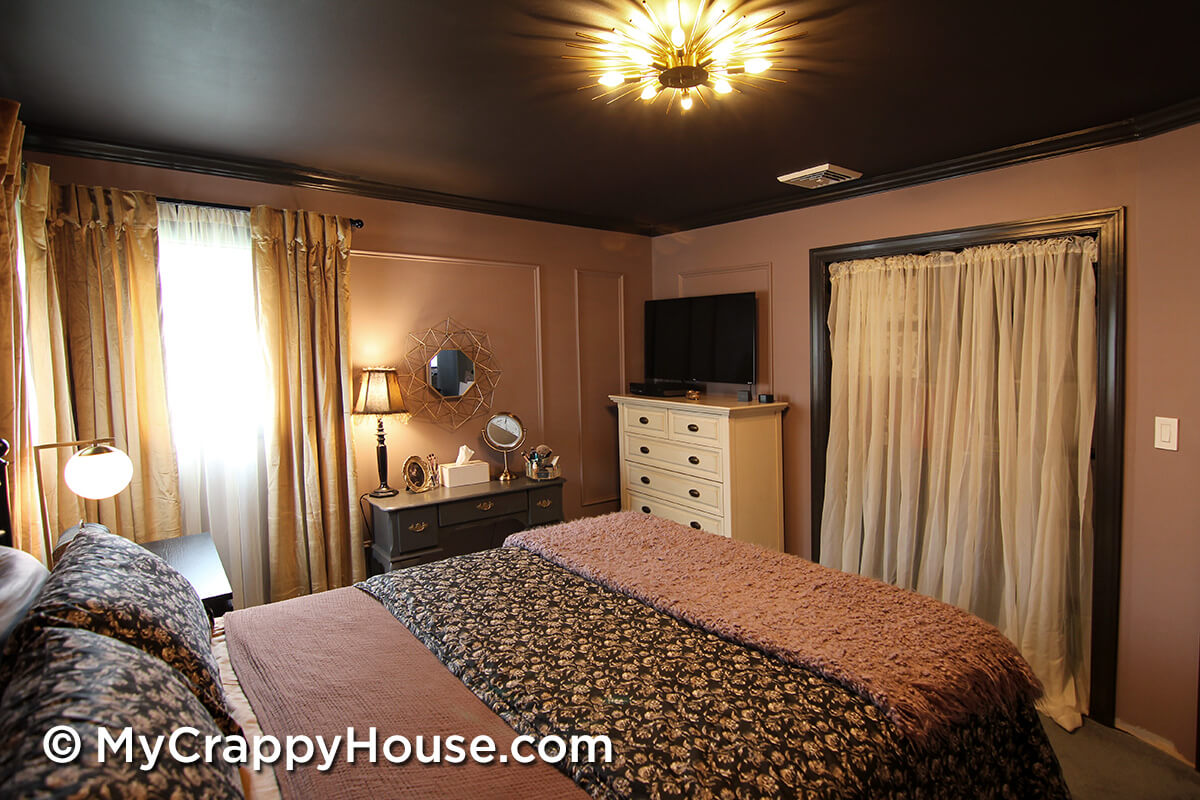 Finished bedroom from reverse angle showing bed with layered bedding, dark painted ceiling with gold starburst light, mauve paneled walls, dresser with TV, and curtained closet.