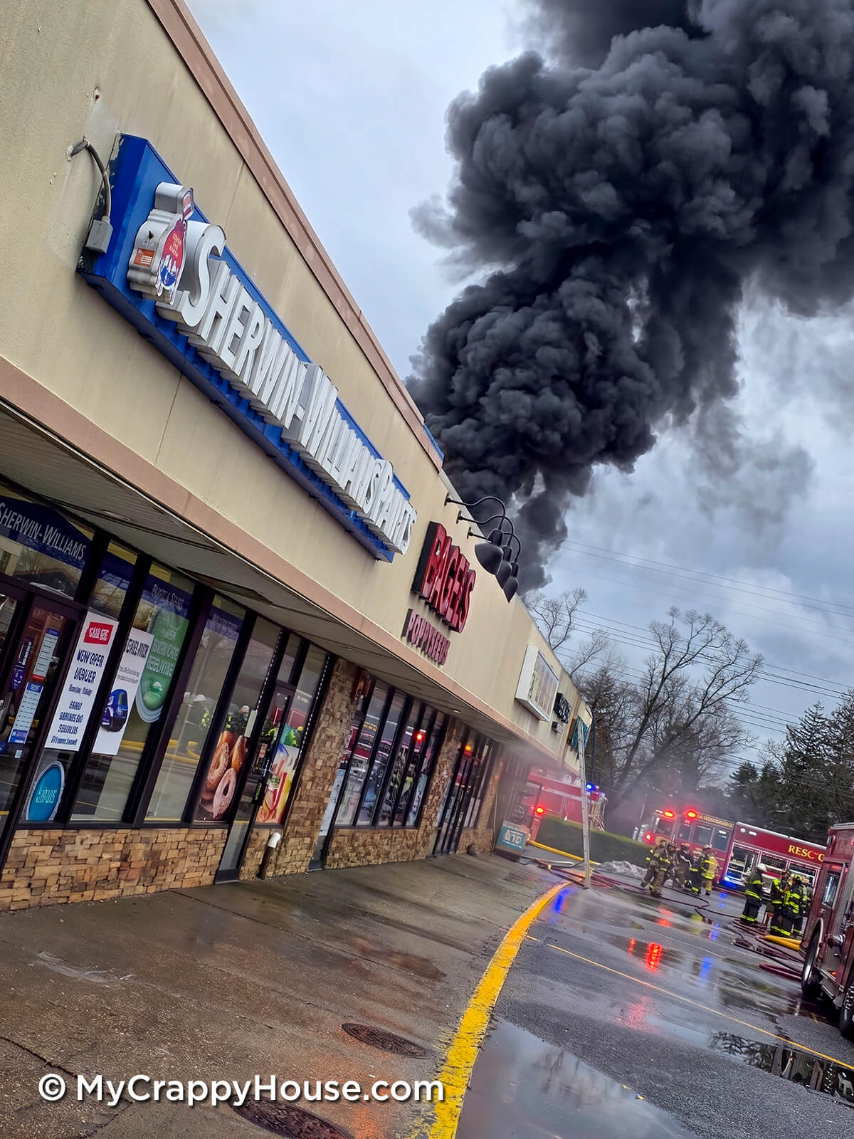 Shopping plaza with a Sherwin-Williams store as thick black smoke billows into the sky while firefighters respond with trucks and hoses.