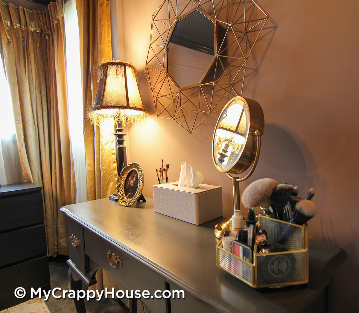 Vanity vignette with gold-framed mirror, lit table lamp, makeup organizer, and soft mauve wall with decorative panel molding in finished bedroom.