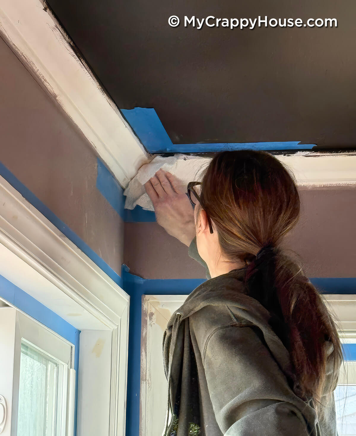 Person wiping and smoothing patched crown molding seam near ceiling corner with painter’s tape still in place during DIY painting and finishing work