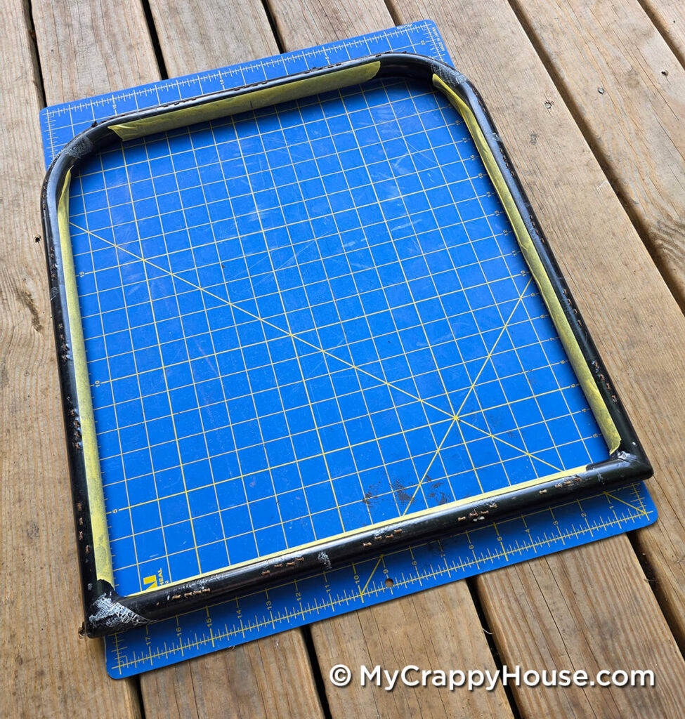 Bare metal chair seat frame on a gridded cutting mat, with masking tape along the inside edges.