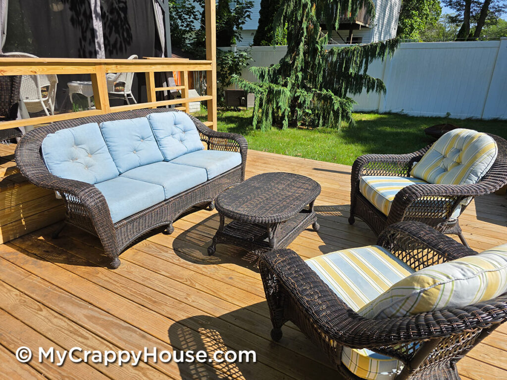 Wicker sofa with blue cushions, two chairs with striped cushions, and a coffee table on a wooden deck before repairs.