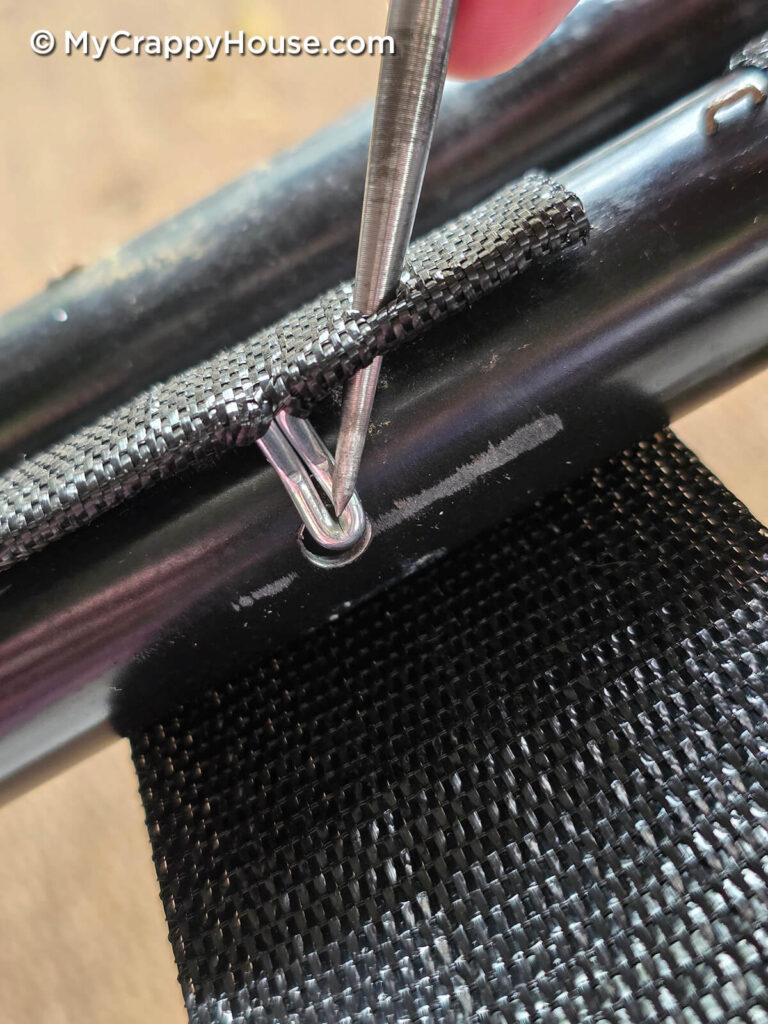 Close-up of an awl passing through black webbing and resting inside the clip’s curved tip at a hole in the metal frame.