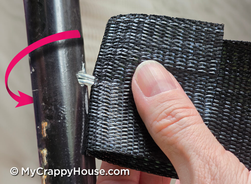 Hand positioning the metal webbing clip at a hole in the chair frame. A pink arrow shows which way to wrap the webbing strip.