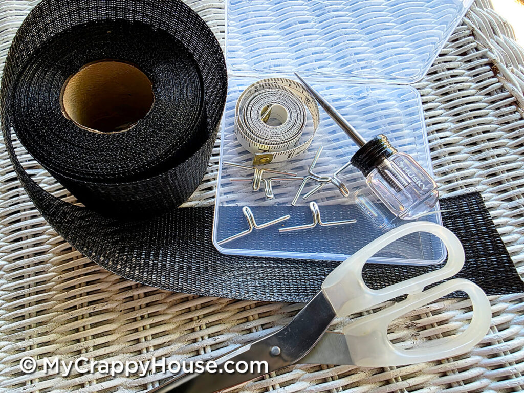 Roll of black replacement webbing, metal clips, an awl, a measuring tape, and scissors arranged on a table.
