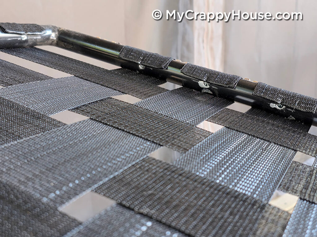 Close-up of black replacement webbing woven in an alternating over-and-under pattern, wrapped around the metal chair frame and secured with clips.